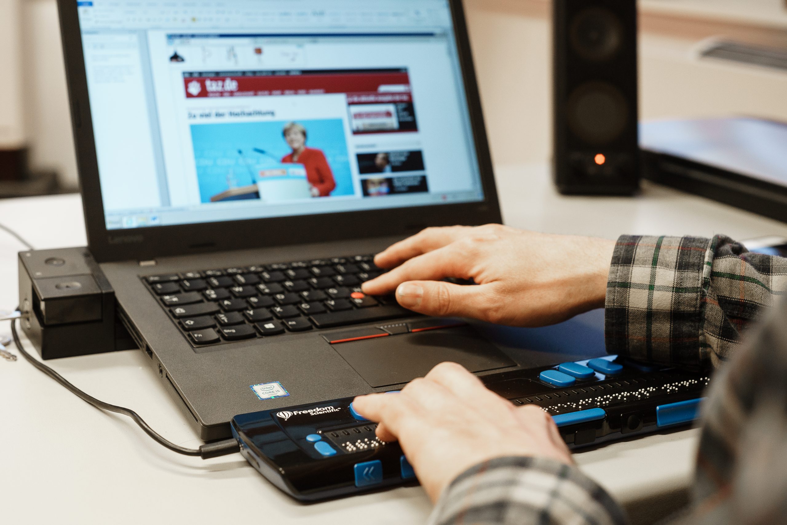Zwei weiße Hände tippen auf die Tastaturen von einem Laptop. Aufgerufen ist eine Internetseite der taz mit einem Foto von Angela Merkel. Die Hände berühren sowohl, die Tastatur des Laptops, als auch eine zusätzlich angeschlossene Tastur für Menschen mit Sehbeeinträchtigung