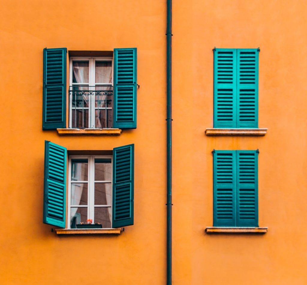 Hausfassade in orange mit zwei geschlossenen und zwei geöffneten Fensterläden in Türkis